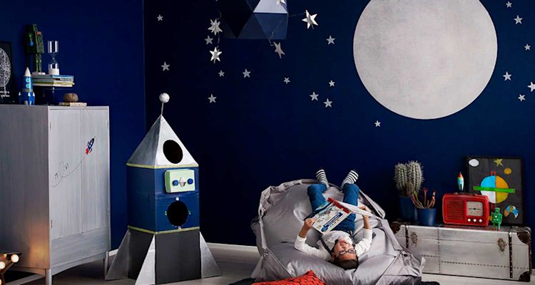 Tummansininen avaruusteemainen huone pahviraketilla, harmaalla säkkituolilla ja tähtitarroilla.