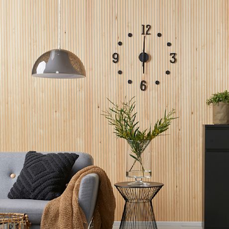 Vaalea puupaneeliseinä, musta minimalistinen kello, harmaa sohva, musta metallipöytä ja vihreä oksa.