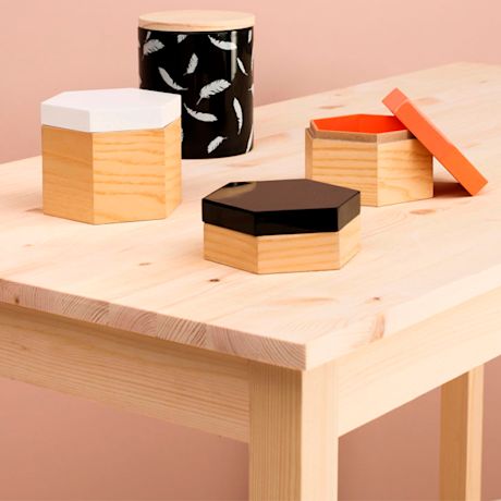 Vaalean puupöydän päällä 3 kuusikulmaista rasiaa eri värisin kansin ja musta sulkakuviopurnukka.