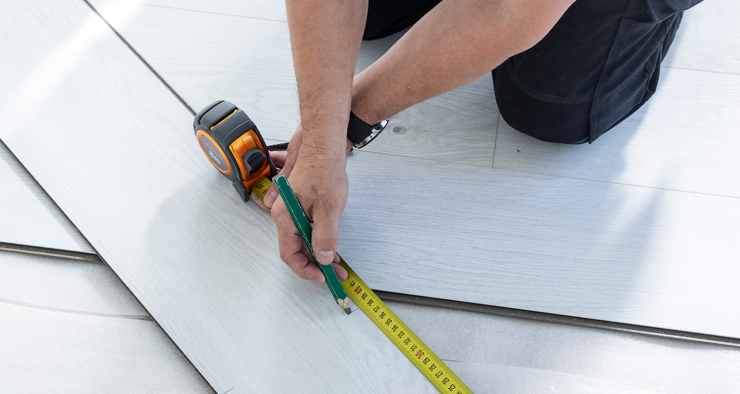 Miehen kädet merkitsevät vaalean lankkulattian mittanauhalla ja vihreällä kynällä.