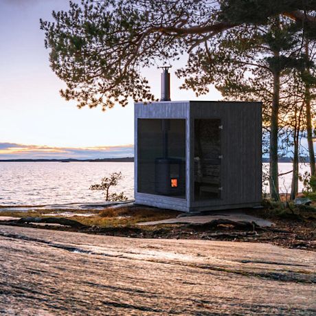 Moderni harmaa saunamökki järven rannalla iltahämärässä, ikkunoista loimuaa oranssi valo.