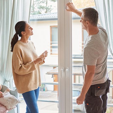 Mies näyttää valkoista lasiparvekeovea beigepuseroiselle naiselle vaaleassa huoneessa.