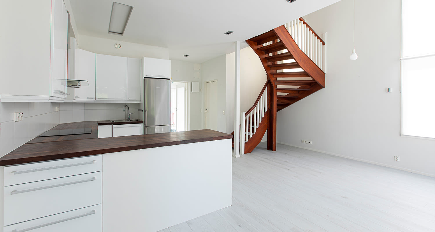 Tyhjä valoisa huone, valkoinen keittiösaareke ja ruskea puuportaikko