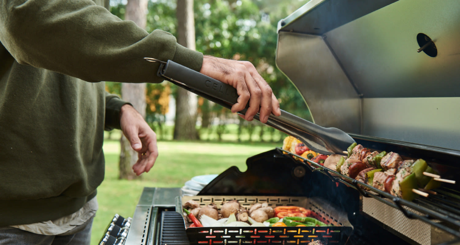 Vihreäpuseroinen käsi grillaa metalligrillissä värikkäitä lihavartaita ja kasviksia ulkoilmassa.