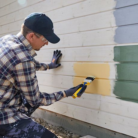 Henkilö maalaa ulkoverhouslautaa keltaisella, vieressä näkyy harmaa ja vihreä värikoelakka.