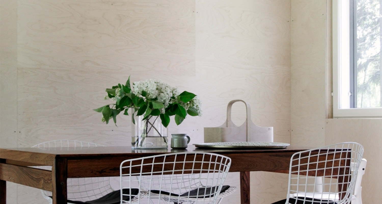 Tumma puupöytä, valkoiset metalliverkkotuolit ja maljakko valkoisia kieloja vaaleassa huoneessa.