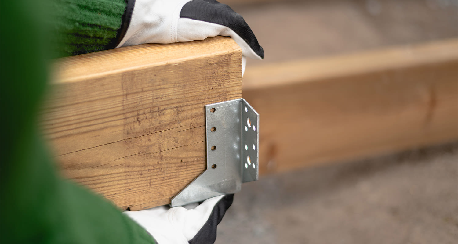 Valko-mustakäsineet nostavat ruskeaa puupalkkia, jossa sinkitty kulmakiinnike.