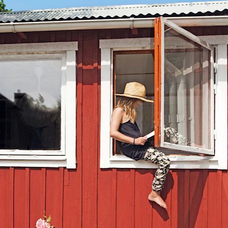 Punaisen mökin avoin ikkunalaudalla istuva lierihattuinen tyttö lukee kirjaa.