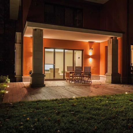 Oranssi talon terassi yöllä, puupöytä tuoleineen liukuovien edessä, nurmikkovalo.