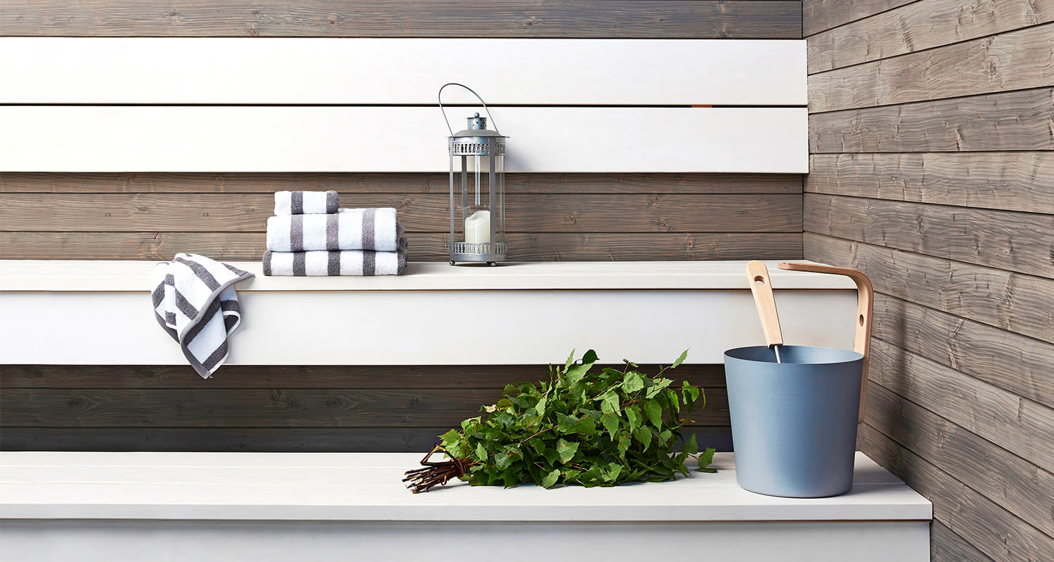 Valkoinen penkki harmaa-valkoisin pyyhkein, hopeinen lyhty, vihreä vasta ja siniharmaa kiulu.