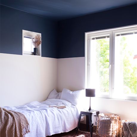 Valoisa makuuhuone: tummansinivalkoinen seinä, valkoinen sänky, pöytälamppu ja korikorit ikkunan edessä