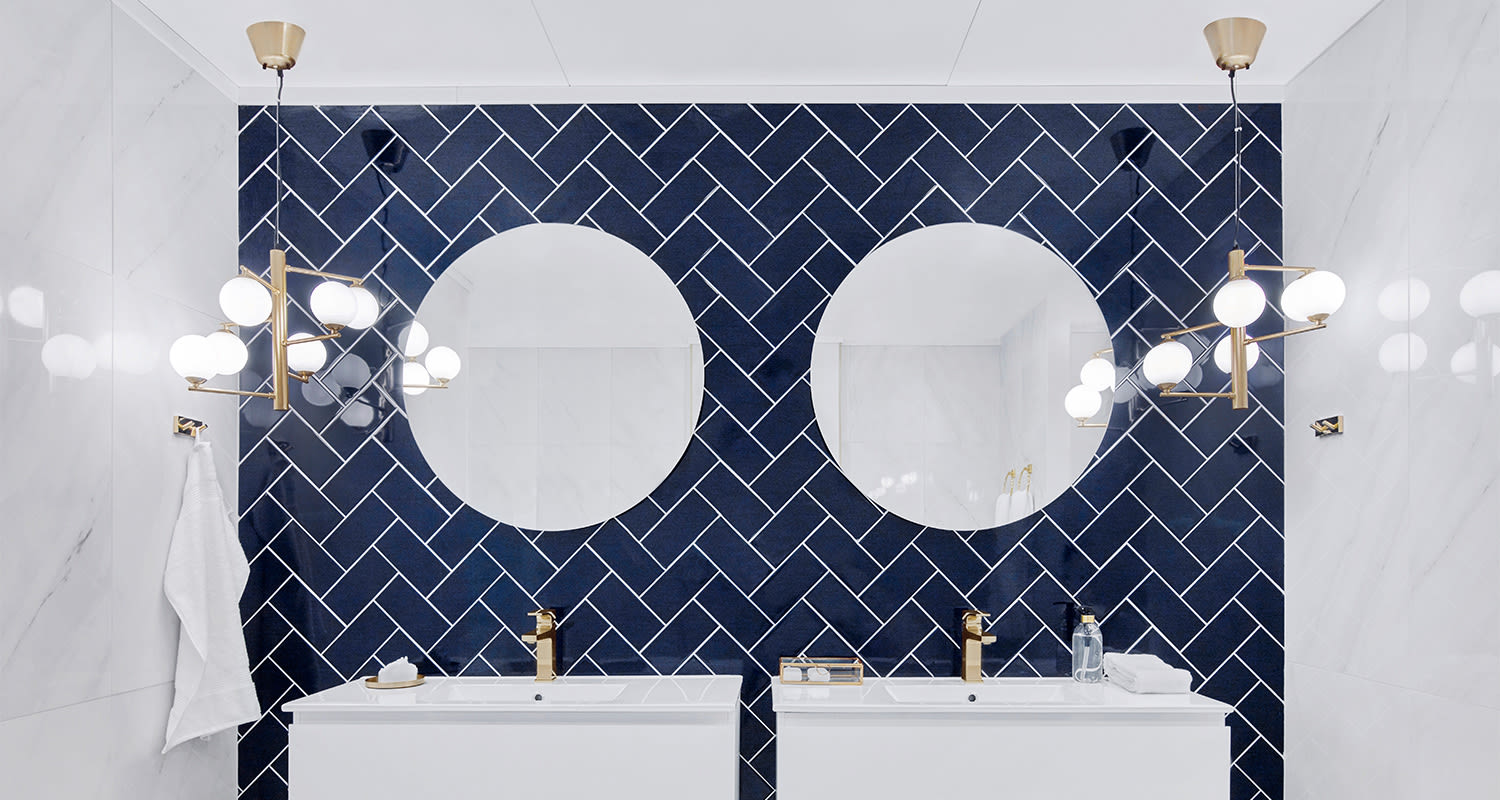 Valkoinen kylpyhuone, sininen kalanruotolaattaseinä, kaksi pyöreää peiliä ja kullanväriset hanat.