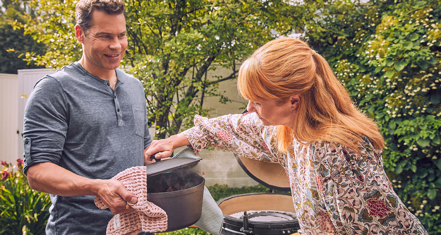 Pariskunta avaa höyryävää grilliä vehreässä takapihassa, mies harmaa paita, nainen kukkapusero.