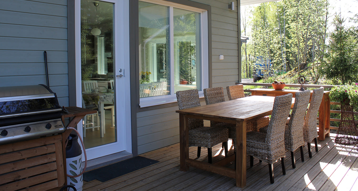 Terassilla puupöytä kuudella punotulla tuolilla, kaasugrilli ja vaalea talon julkisivu.