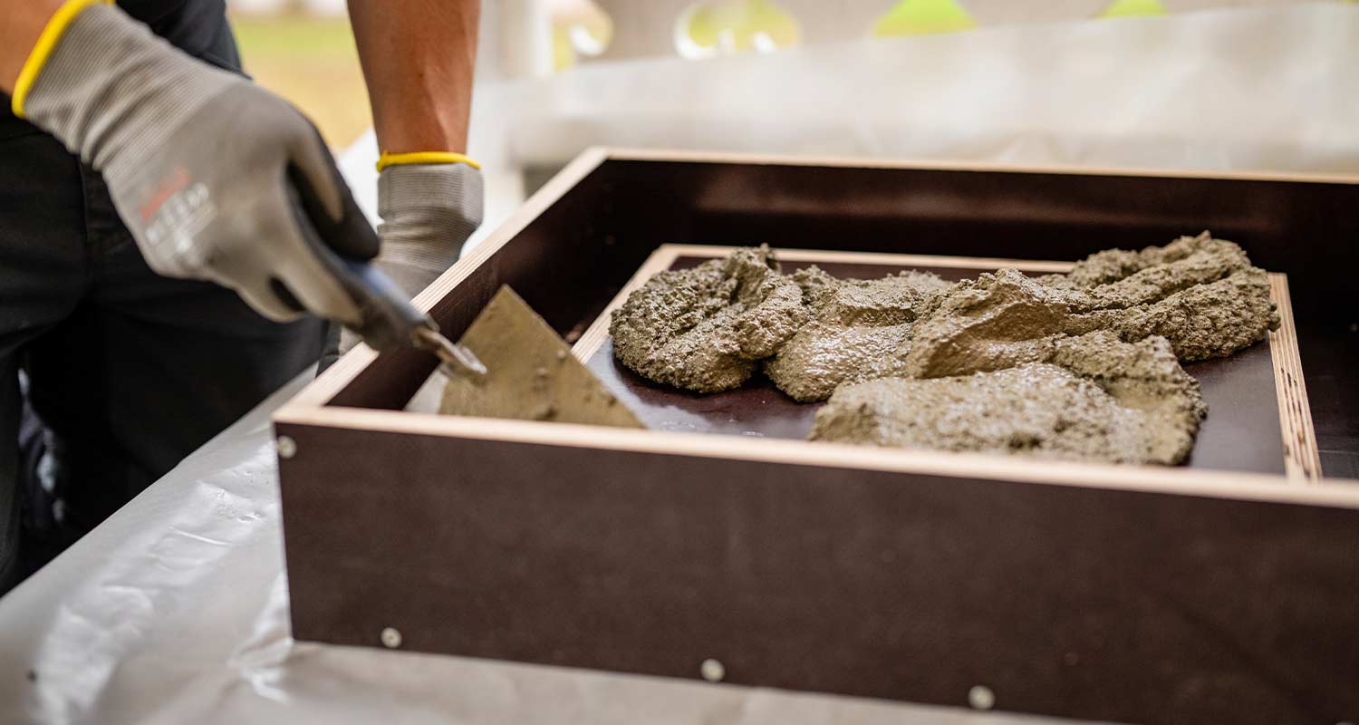 Rakennustyöläinen levittää lastalla harmaata laastia ruskeaan muottiin suojahanskoissa