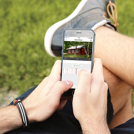 Kädet pitelevät puhelinta vihreällä nurmella, ruudulla valvontakameran tallenne punaisesta mökistä