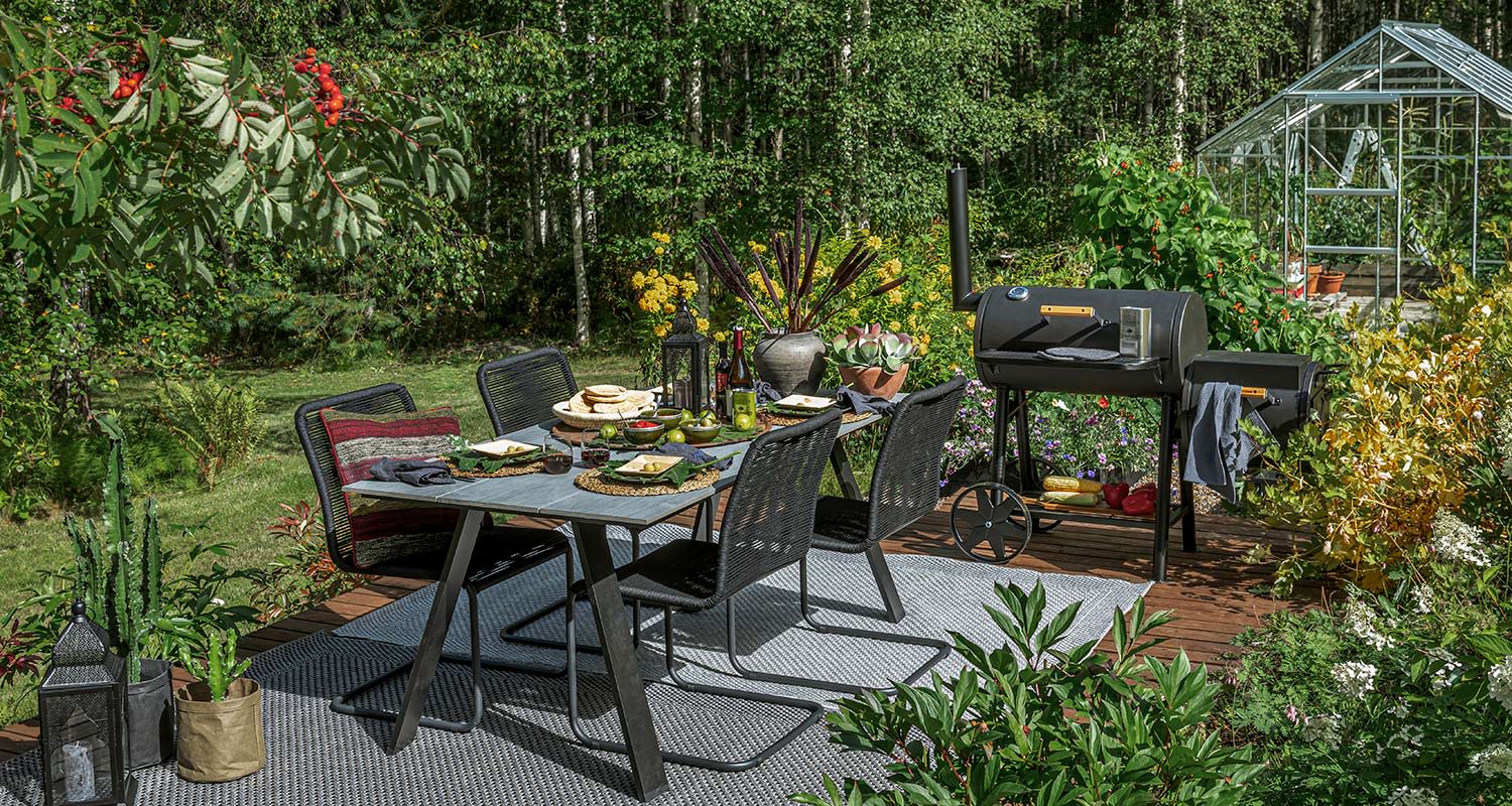 Pihaterassilla mustat tuolit ympäröivät katettua ruokapöytää, grilli ja kasvihuone taustalla.