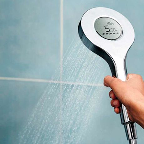 Käsi pitää pyöreää hopeista suihkupäätä diginäytöllä, josta valuu vettä sinistä laattaa vasten.