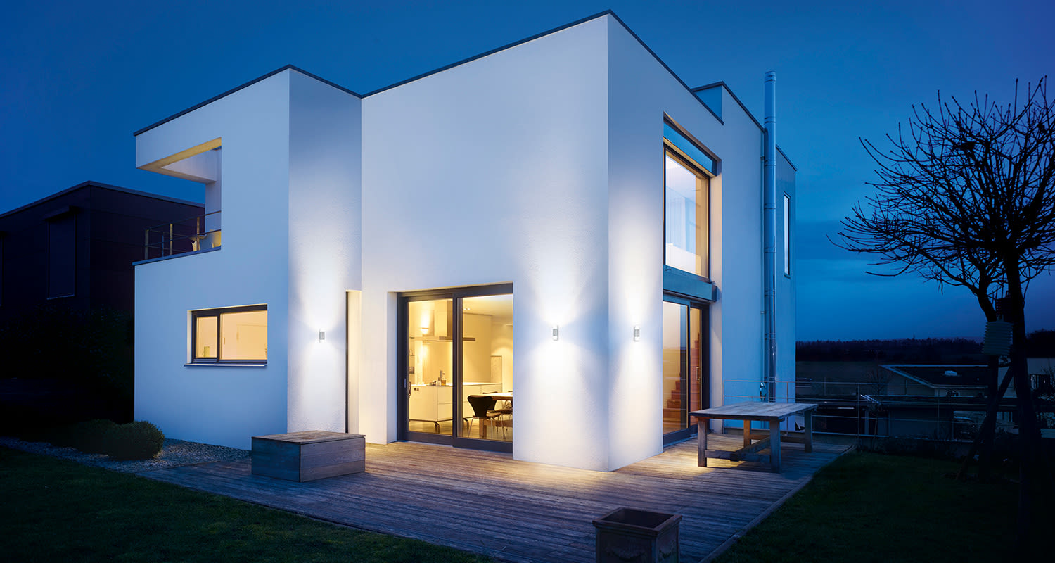 Valkoinen kaksikerroksinen talo puulautaterassilla illan sinisellä taivaalla, ikkunavalo.