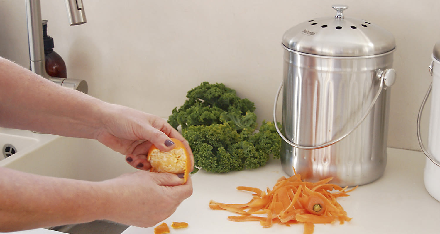 Kädet kuorivat appelsiinia keittiössä, vieressä vihreää lehtikaalia ja metallinen kompostiastia