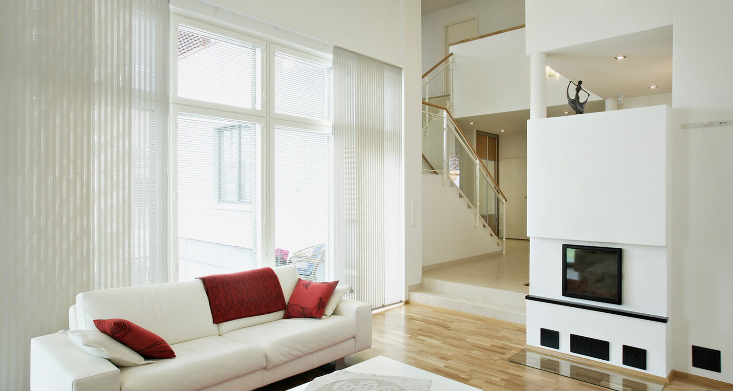 Moderni valkoinen olohuone, sohva punaisin tyynyin ja vilttipeitteellä suuren ikkunan vieressä.