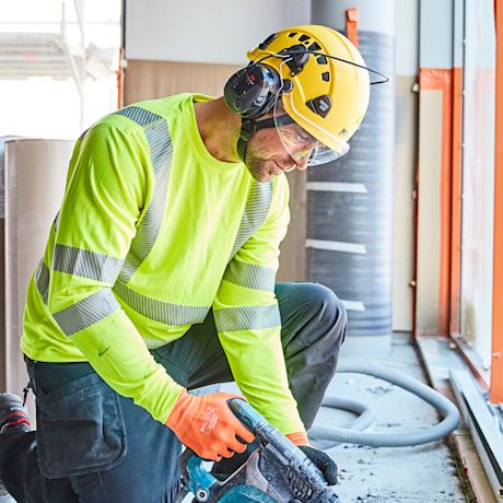 Rakennustyömies neonkeltaisessa paidassa ja keltaisessa kypärässä kyykyssä käyttää sähkötyökalua.