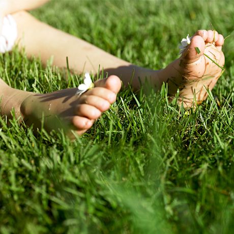 Lapsen paljaat jalat kesäisellä ruohikolla, ruohoa varpaissa ja pieni valkoinen kukka.