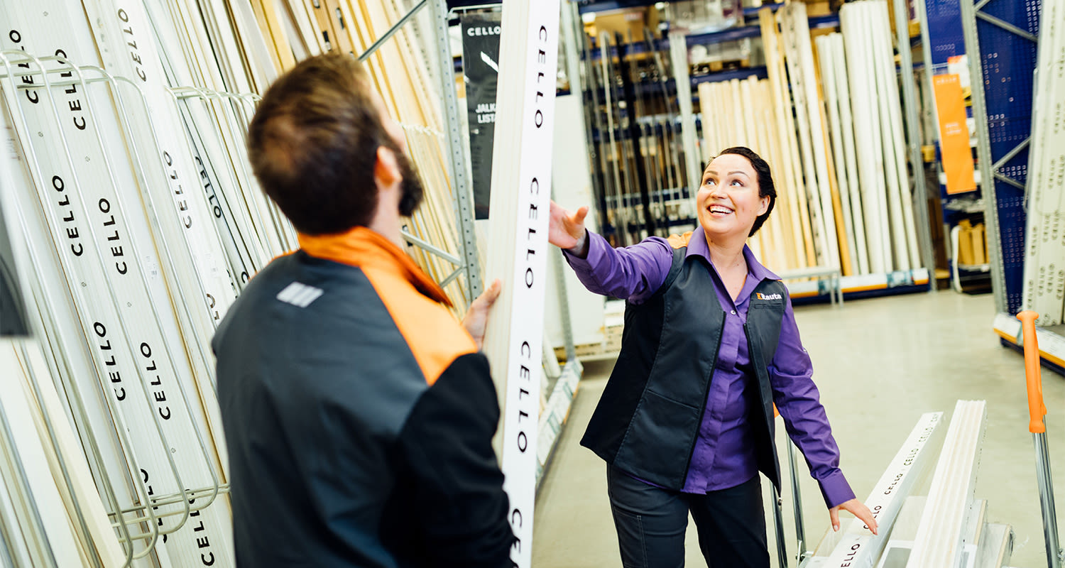 Kaksi rautakaupan työntekijää ojentaa valkoista CELLO-karmilistaa hyllyjen edessä.