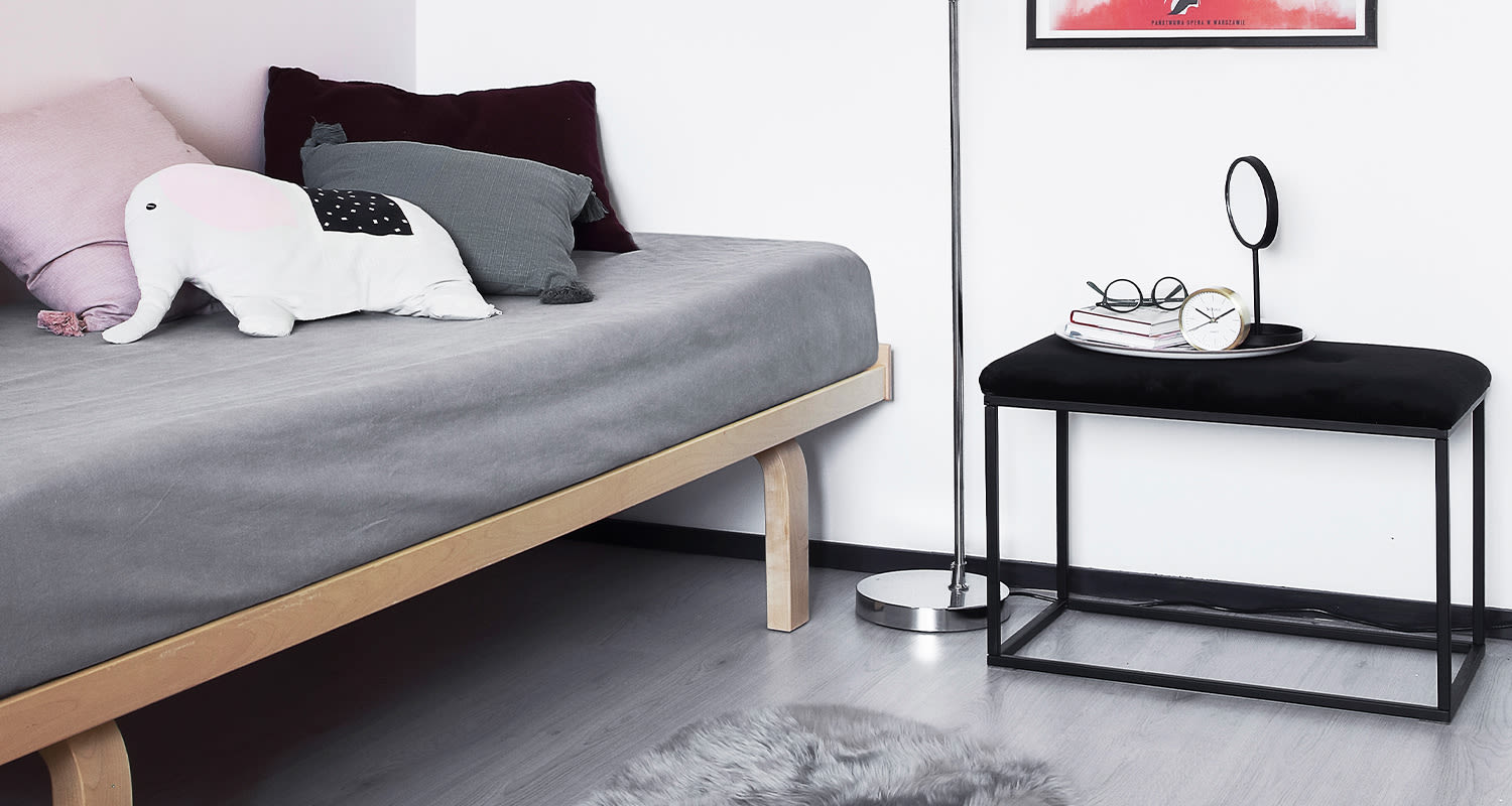 Vaalean puun harmaa sänky pastellityynyillä ja norsutyynyllä, musta metallipenkki kirjoineen.