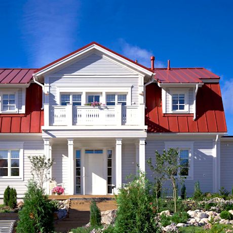 Valkoinen puutalo punaisella katolla, parveke kukkineen ja vehreä etupiha.