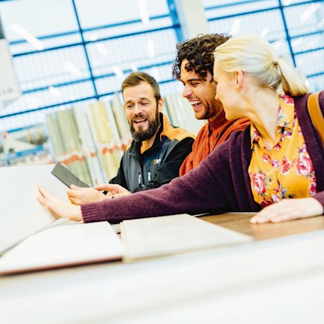 Kolme ihmistä vertailevat laattanäytteitä kirkkaassa rakennustarvikemyymälässä.