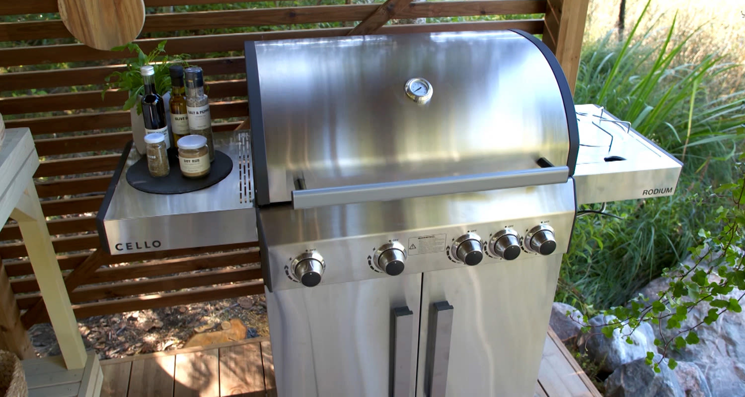 Hopeanhohtoinen kaasugrilli ulkoterassilla, viisi säätönuppia, sivuhyllyllä yrttejä ja mausteita