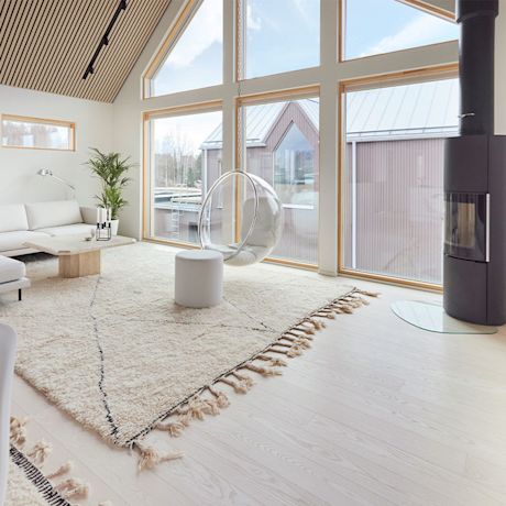 Vaalea olohuone suurilla ikkunoilla, roikkuva läpinäkyvä tuoli, beige matto ja musta takka