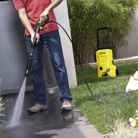 Punapaitainen mies pesee terassia keltaisella painepesurilla, ruohoa ja pensaita.