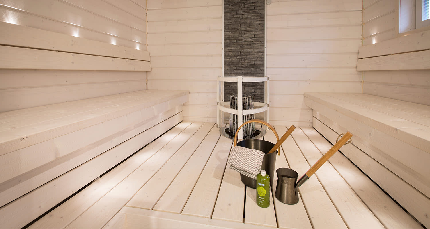 Vaalea moderni puusauna, keskellä kiuas, lattiolla musta löylykiulu ja vihreä tervapullo.