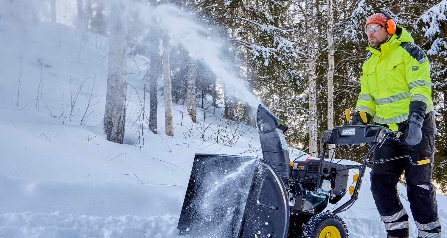 Henkilö keltaisessa huomiotakissa työntää lumikonetta metsäisellä lumisella tiellä.