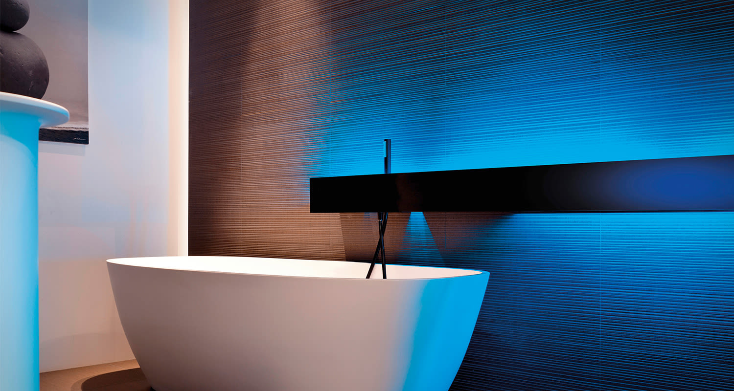 Valkoinen kylpyamme ruskean tekstuuriseinän edessä sinisellä LED-valaistuksella