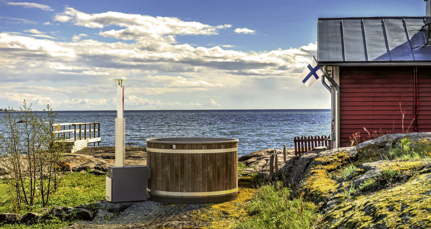 Punainen mökki kalliorannalla, puinen kylpytynnyri, laituri ja Suomen lippu.