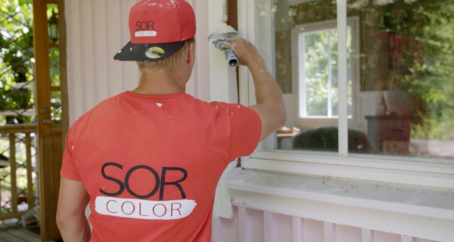 Rakennusmaalari punaisessa SOR Color-paidassa ja ‑lippiksessä maalaa valkoista ikkunapuitetta.