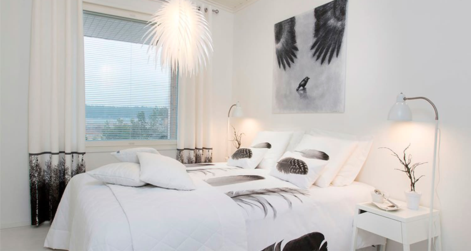 Vaalea makuuhuone, valkoinen sänky mustavalkoisilla sulkakuviotekstiileillä ja seinätaulu.