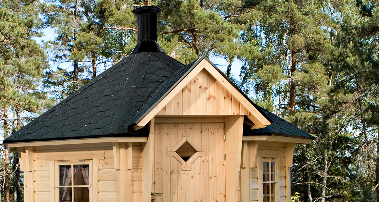 Vaaleasta puusta rakennettu grillimaja mustalla huopakatolla havumetsän keskellä.