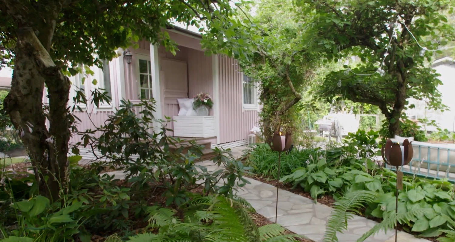 Vaaleanpunainen puutalo, vihreä köynnöskaari ja puutarha kivilaattapolulla