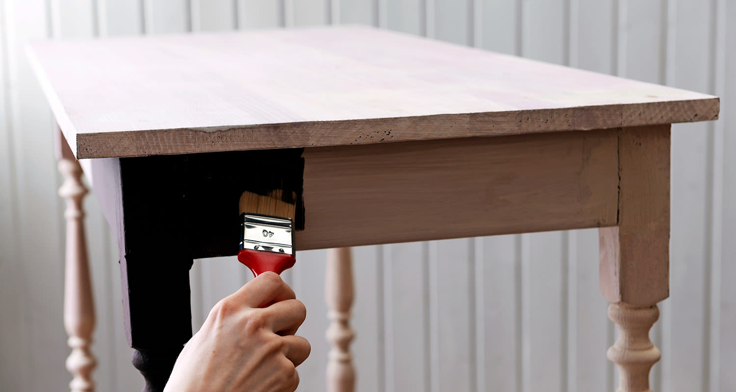 Käsi pitelee punaista sivellintä ja maalaa vaaleaa puupöytää mustalla maalilla.