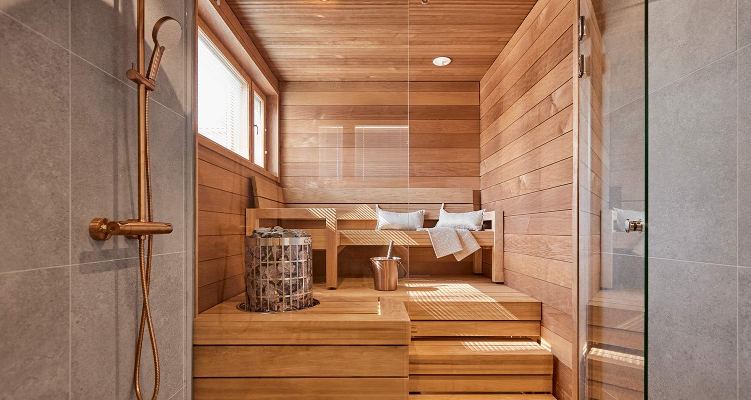 Vaalea puusauna harmaalla laatoitetulla suihkuseinällä, puiset penkit, kiuaskori ja kuparikauha.