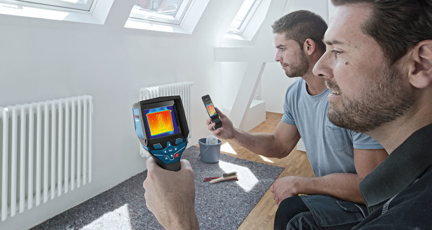 Kaksi asentajaa ullakkohuoneessa mittaa patterin lämpökuvaa termokameralla ja älypuhelimella.