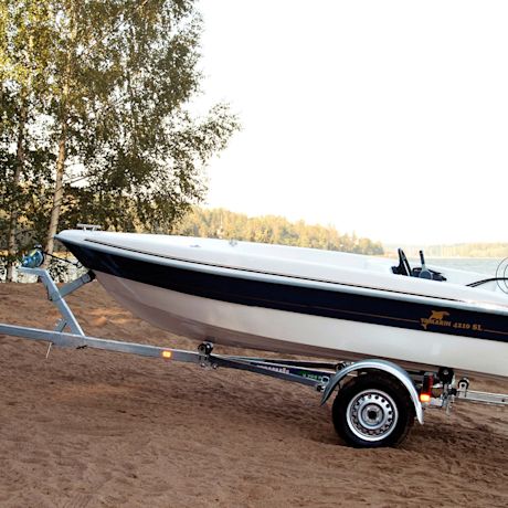 Valkoinen-tummansininen moottorivene kärryssä järven hiekkarannalla koivun juurella