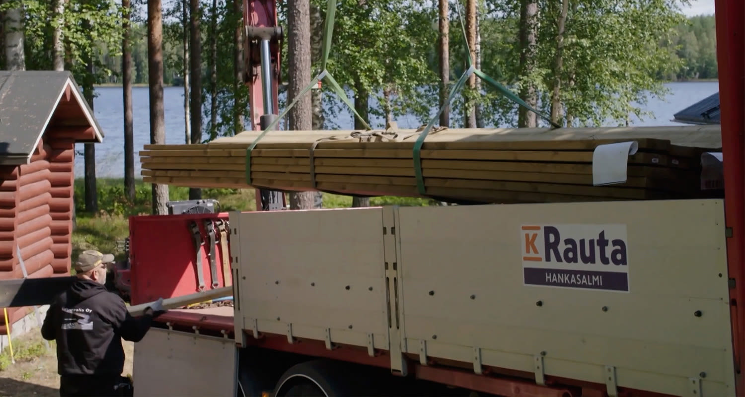 Punainen K-Rauta Hankasalmi -kuorma-auto purkaa vaaleita lankkuja järvenrantamökin luona.