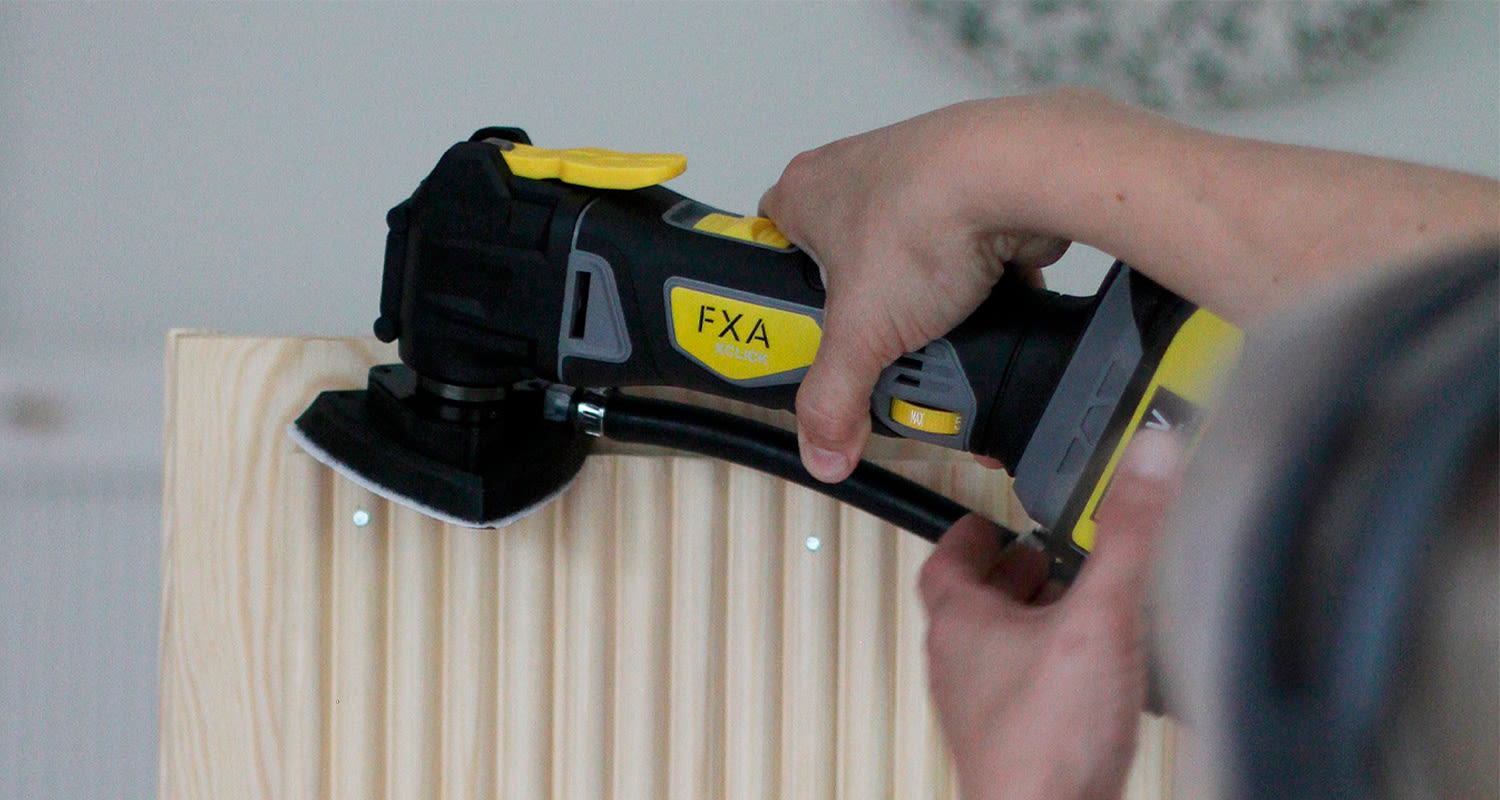 Käden käyttämä musta-keltainen FXA XClick -monitoimityökalu hioo vaaleaa puulevyä.