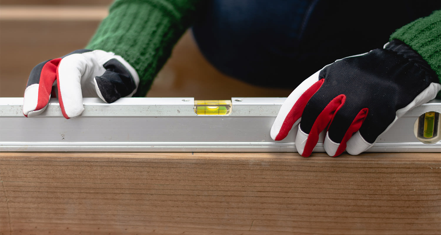 Punamustavalkoiset työhanskat pitävät hopeista vesivaakaa vaakasuorassa puupalkin päällä.