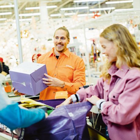 Oranssipaitainen myyjä skannaa violettia pakettia rautakaupassa, asiakas pakkaa liilaan kassiin.